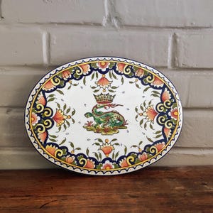 Peut inclure: Assiette ovale en céramique à fond blanc, ornée d'un motif central représentant un dragon vert couronné. L'assiette est bordée d'un motif floral orange, jaune et bleu. L'assiette est présentée sur une surface en bois.