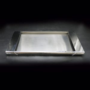 Könnte beinhalten: Ein rechteckiges Tablett aus Edelstahl mit gebürsteter Metalloberfläche. Das Tablett hat ein niedriges Profil mit leicht erhöhten, abgewinkelten Seiten an jedem Ende. Das Design ist minimalistisch und modern.