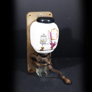Puede incluir: Antiguo molinillo de café con un recipiente de cerámica blanca con una ilustración caprichosa de una niña con una jaula de pájaros. El molinillo está montado sobre una tabla de madera y tiene un mecanismo de molienda de metal y un tarro de recogida de vidrio.