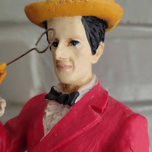 May include: A decorative figurine of a man in a red suit, black bow tie, and yellow top hat, holding a monocle. The figure has black hair and blue eyes. A collectible item.