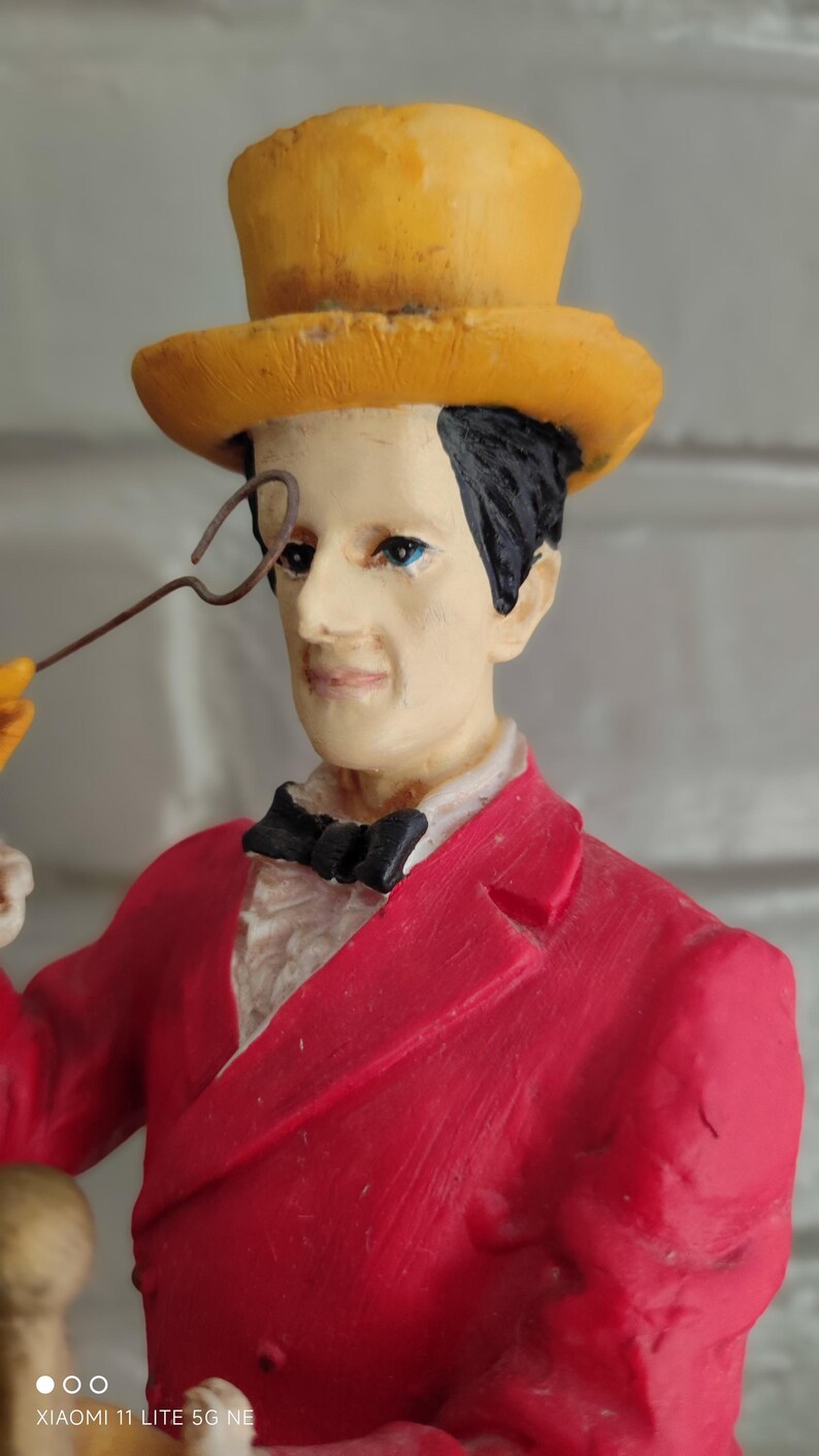 May include: A decorative figurine of a man in a red suit, black bow tie, and yellow top hat, holding a monocle. The figure has black hair and blue eyes. A collectible item.