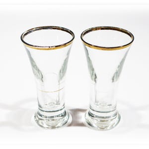 Peut inclure: Deux verres à boire en verre transparent avec un bord doré. Les verres ont une forme évasée et une petite base. Les verres sont vides et reposent sur une surface blanche.