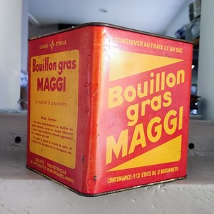 Peut inclure: Une boîte en métal rouge et jaune avec le texte "Bouillon gras MAGGI" dessus. La boîte est étiquetée avec des instructions pour la conserver au frais et au sec. La boîte contient 112 cubes de bouillon.