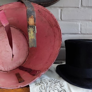 May include: A black top hat positioned next to a large, round, open container with a pink interior lining. The container features worn leather straps attached to the inner walls, showing signs of age and use with visible scuffs and discoloration.