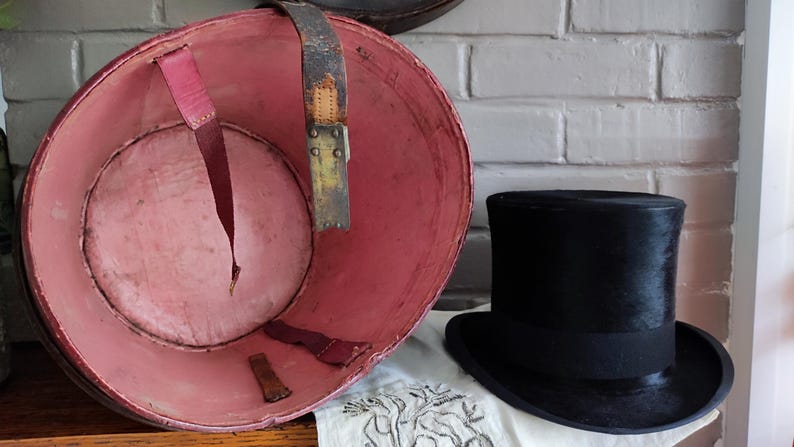 May include: A black top hat positioned next to a large, round, open container with a pink interior lining. The container features worn leather straps attached to the inner walls, showing signs of age and use with visible scuffs and discoloration.