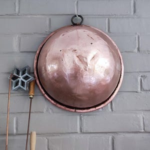 Peut inclure: Un grand bol rond en cuivre poli avec une surface lisse et réfléchissante, doté d'un anneau de suspension en métal sombre sur le dessus. À côté, un moule en métal en forme d'étoile est fixé à une longue tige en cuivre avec un manche en bois clair.