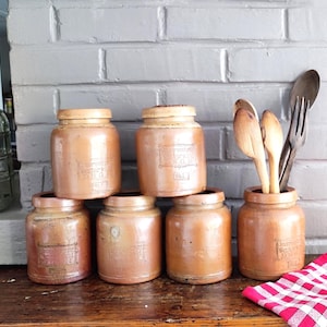 May include: A collection of six cylindrical tan ceramic storage jars with lids, featuring embossed rectangular labels. One jar contains three wooden spoons and a wooden fork. A red and white checkered cloth is placed next to the jars.