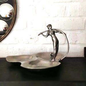 May include: A silver-toned metal serving dish featuring a three-sectioned tray base and a central sculptural figure of a person standing while holding a curved handle that arches over the compartments, creating a decorative and functional centerpiece.