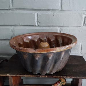 May include: An antique brown and black bundt pan with a central tube, resting on a small wooden table. The pan has a fluted design and a light brown rim. The background is a white brick wall.
