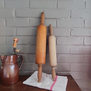 Peut inclure: Deux rouleaux à pâtisserie en bois de différentes tailles se dressent contre un mur de briques grises. Le plus grand est brun clair chaud, tandis que le plus petit est de couleur bois naturel plus clair. Un pichet marron et une horloge sont également visibles.