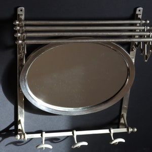May include: A silver wall-mounted mirror with a rounded oval shape and three hooks below. The mirror has a silver frame and is attached to a silver wall bracket.