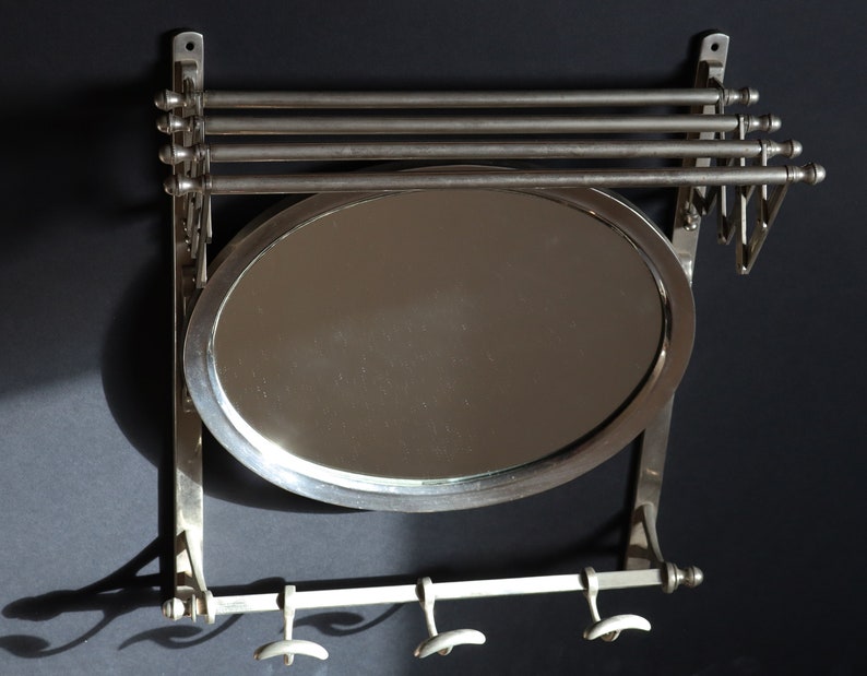 May include: A silver wall-mounted mirror with a rounded oval shape and three hooks below. The mirror has a silver frame and is attached to a silver wall bracket.