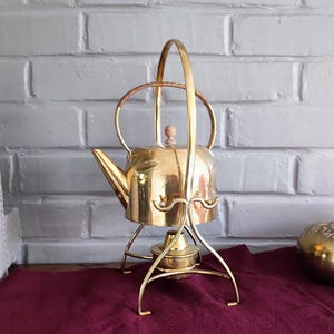 May include: A vintage brass tea kettle with a curved handle and a stand. The kettle has a spout and a wooden knob on the lid. The stand has a burner base. The kettle is sitting on a burgundy cloth.