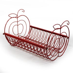 Può includere: Un cesto portafrutta in filo metallico rosso con corpo cilindrico a doghe aperte e due pannelli terminali decorativi a forma di contorno di mela, ciascuno sormontato da un piccolo dettaglio a stelo e foglia, realizzato in sottili aste.