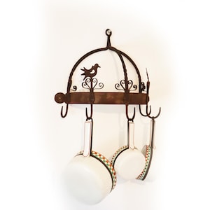 May include: A wrought iron pot rack with a decorative bird silhouette and scrollwork. Three white pots with red, green, and white patterned rims hang from the rack. The rack is mounted on a white wall.