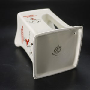 May include: White ceramic ashtray with a rectangular base and a handle. The ashtray features the text "Johnnie Walker Scotch Whisky" in red and black, along with a graphic of a man walking. The bottom has a logo and text.