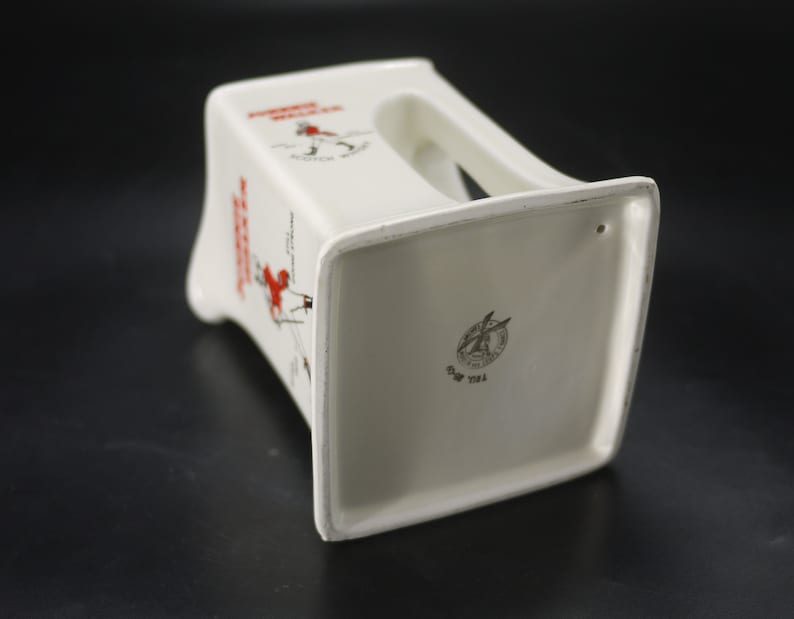 May include: White ceramic ashtray with a rectangular base and a handle. The ashtray features the text "Johnnie Walker Scotch Whisky" in red and black, along with a graphic of a man walking. The bottom has a logo and text.