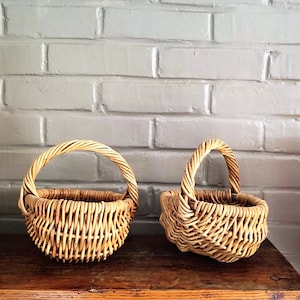 May include: Two small, round, light brown woven wicker baskets with arched handles. Each basket features a sturdy, open-weave construction made from natural plant fibres, showcasing a textured, interlaced pattern throughout the body and handle.