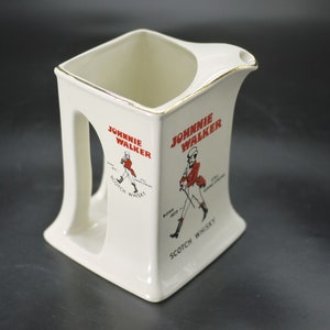 May include: Cream-coloured ceramic pitcher with a square base and a handle. The pitcher features the "Johnnie Walker" logo in red, with an illustration of the Striding Man. The rim is trimmed with gold. "Scotch Whisky" is printed below the logo.