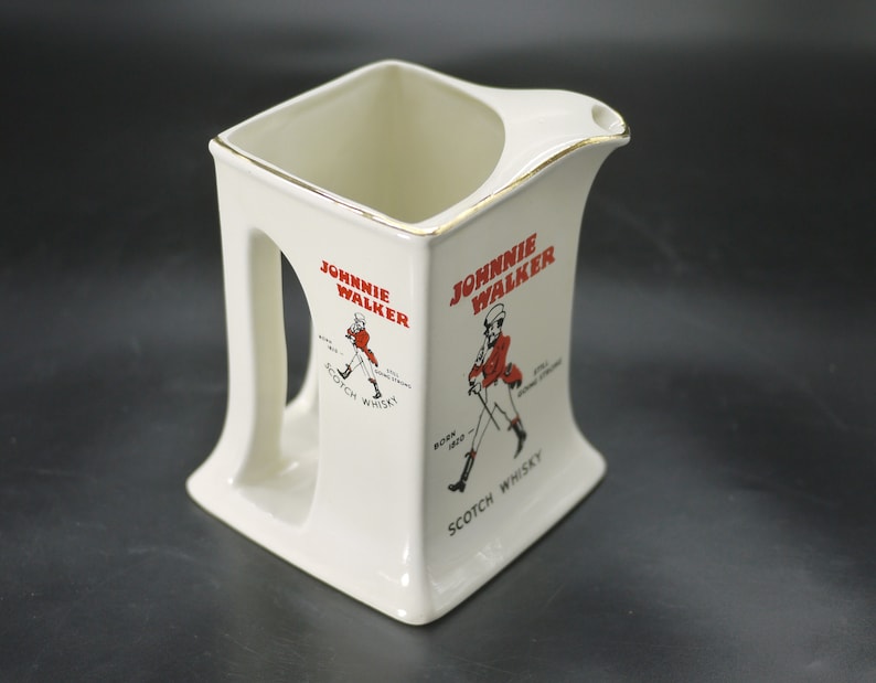 May include: Cream-colored ceramic pitcher with a square base and a handle. The pitcher features the "Johnnie Walker" logo in red, with an illustration of the Striding Man. The rim is trimmed with gold. "Scotch Whisky" is printed below the logo.