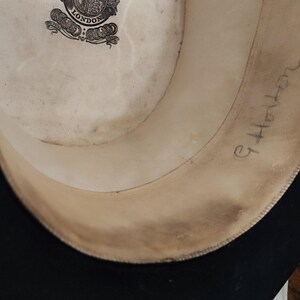 May include: The interior of a black hat featuring a cream-colored lining with a circular logo that reads London. The inner band is made of a light tan material and has the handwritten name G. Horton written in cursive ink on the right side.