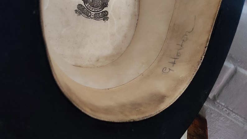 May include: The interior of a black hat featuring a cream-colored lining with a circular logo that reads London. The inner band is made of a light tan material and has the handwritten name G. Horton written in cursive ink on the right side.