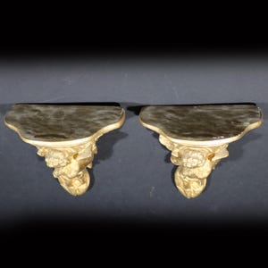 May include: A pair of gold-painted wooden wall shelves with ornate floral and scrollwork designs. The shelves are shaped like a crescent moon and have a slightly curved top edge.