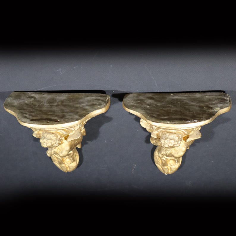 May include: A pair of gold-painted wooden wall shelves with ornate floral and scrollwork designs. The shelves are shaped like a crescent moon and have a slightly curved top edge.