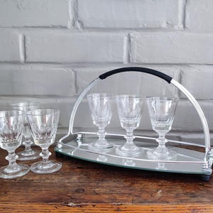May include: A set of clear crystal cordial glasses with a silver-toned serving tray. The tray has a handle with a black grip and holds four glasses. Additional glasses are placed to the left of the tray on a wooden surface.