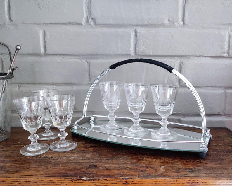 May include: A set of clear crystal cordial glasses with a silver-toned serving tray. The tray has a handle with a black grip and holds four glasses. Additional glasses are placed to the left of the tray on a wooden surface.