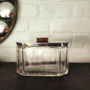 May include: A clear glass container with a faceted, hexagonal shape and a metallic silver-toned rim. The lid features a small, rectangular dark brown wooden handle centered on top, providing a contrast to the transparent glass body.