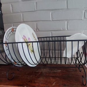 Puede incluir: Un escurridor de platos de alambre de metal negro con marco rectangular y patas decorativas en forma de voluta. El soporte sostiene dos platos de cerámica blanca, uno con un patrón floral y el otro con una ilustración de vegetales.