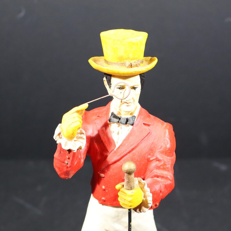 May include: A figurine of a man in a red jacket, yellow top hat, and yellow gloves, holding a monocle and a cane. The figure has a black bow tie and a white shirt. The figurine is a vintage collectible.