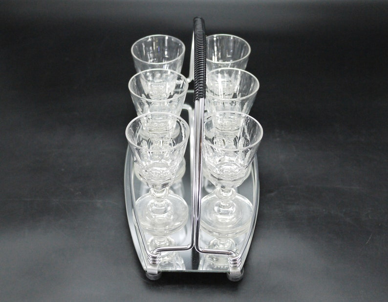 May include: A vintage silver-toned serving tray with a handle, holding six clear crystal stemmed glasses. The glasses have a decorative pattern and are arranged in two rows of three. The handle is black and ribbed.
