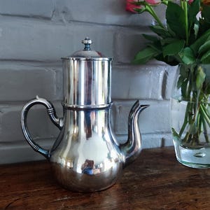 Peut inclure: Une cafetière en métal argenté poli dotée d'une section supérieure cylindrique, d'une base arrondie, d'une anse incurvée et d'un bec verseur fin. La cafetière possède un couvercle amovible avec un petit bouton sur le dessus, reflétant la lumière.