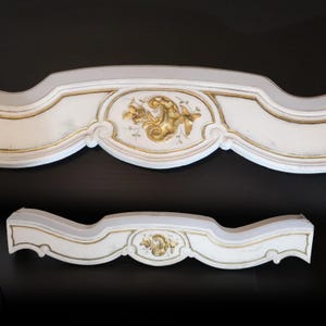 Può includere: Due elementi architettonici decorativi in legno bianco con dettagli dorati. Ogni pezzo presenta un motivo floreale dorato centrale. I pezzi hanno una forma curva e ornata, probabilmente parte di una struttura decorativa più grande.