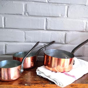 Peut inclure: Trois casseroles en cuivre avec des intérieurs argentés métalliques et de longues poignées en métal sombre. Les casseroles sont disposées en rangée, la plus grande reposant sur un linge blanc plié, révélant leurs surfaces extérieures polies.