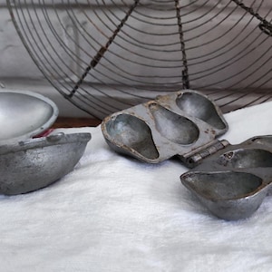 May include: Several vintage metal egg poachers and molds rest on a white towel with a red stripe. The poachers are various shapes and sizes, with a wire cooling rack in the background. The items appear to be antique kitchenware.