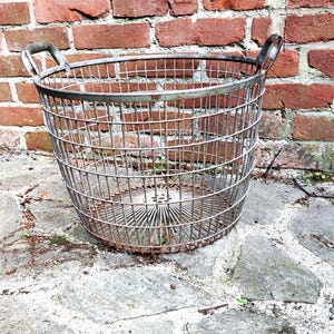 Könnte beinhalten: Ein runder Drahtkorb aus verwittertem Metall mit einer sichtbaren Rostpatina. Der Korb verfügt über einen kreisförmigen Boden mit strahlenförmigen Drahtspeichen, einen verstärkten oberen Rand und zwei geschwungene Metallgriffe an den Seiten.