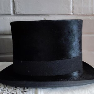 May include: A black top hat with a flat crown and a wide, curved brim. The hat features a textured, dark surface and a solid black band encircling the base of the crown. The structure is cylindrical with a distinct, formal silhouette.