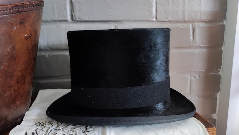 May include: A black top hat with a flat crown and a wide, curved brim. The hat features a textured, dark surface and a solid black band encircling the base of the crown. The structure is cylindrical with a distinct, formal silhouette.