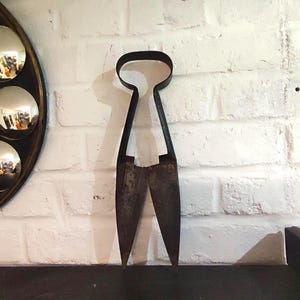 Peut inclure: Une grande cisaille en métal noir antique avec une poignée en boucle, posée contre un mur de briques blanches. Les lames sont ouvertes et la cisaille projette une ombre. Un miroir rond avec plusieurs miroirs convexes est visible à gauche.