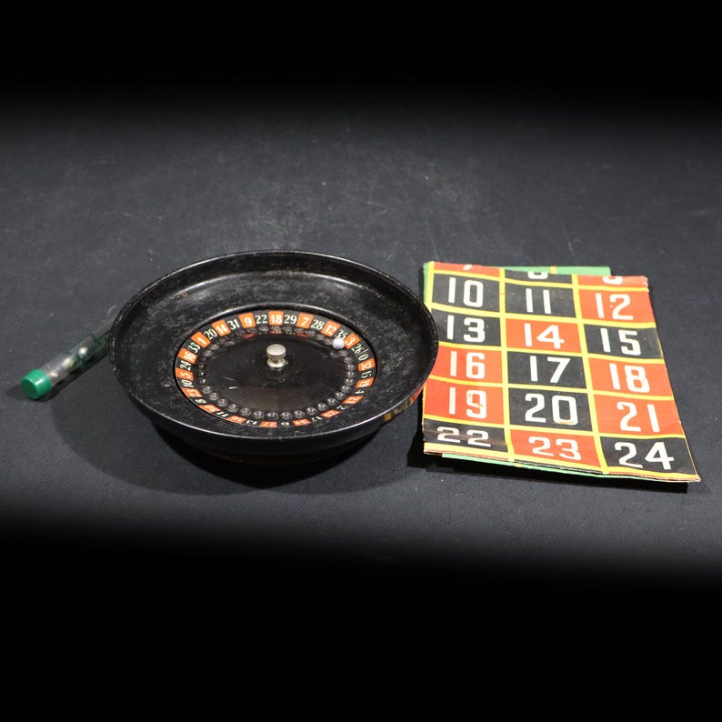 May include: A black roulette wheel with a central silver spindle, a small white ball, and a green plastic tube containing additional balls. Beside the wheel is a rectangular betting mat featuring a grid of numbers from 10 to 24 in white.
