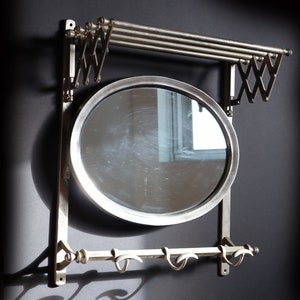 May include: A silver wall-mounted mirror with a round frame and a shelf above it. The shelf has two metal bars that can be extended to hold clothes. There are also three hooks below the mirror.