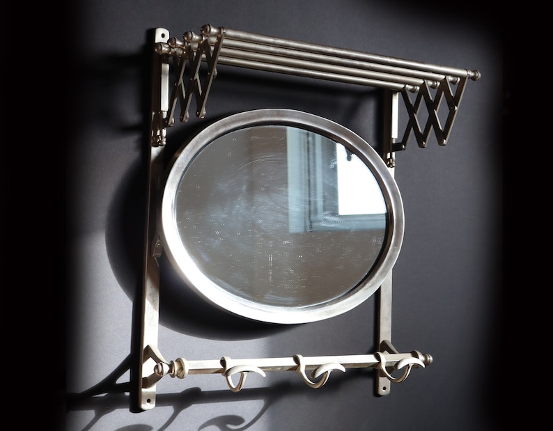 May include: A silver wall-mounted mirror with a round frame and a shelf above it. The shelf has two metal bars that can be extended to hold clothes. There are also three hooks below the mirror.
