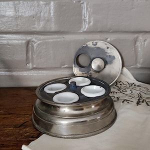 May include: A vintage silver-toned egg holder with four white porcelain egg cups. The lid is off to the side. The egg holder sits on a wooden surface, with a white cloth and brick wall in the background.