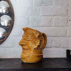 Peut inclure: Pichet en céramique de couleur beige représentant un visage d'homme avec une barbe et des traits détaillés. Il possède une anse et est posé sur une surface sombre devant un mur de briques blanches. Un miroir décoratif est visible en arrière-plan.