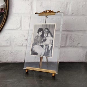 Peut inclure: Un cadre photo en verre transparent avec un support et des accents en métal doré. Le cadre contient une photographie en noir et blanc d'une famille. Le cadre est exposé sur une surface sombre.