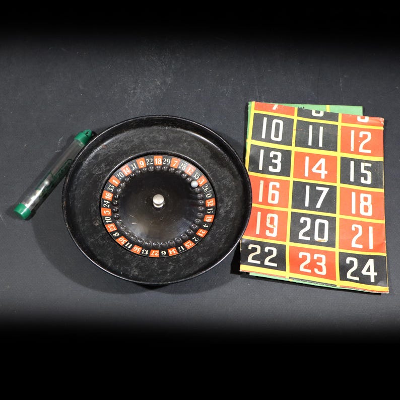 May include: A black roulette wheel with a central silver spindle, a small silver ball, a clear plastic tube with green caps containing additional balls, and a rectangular betting mat featuring a grid of numbers from 10 to 24 in red and black.