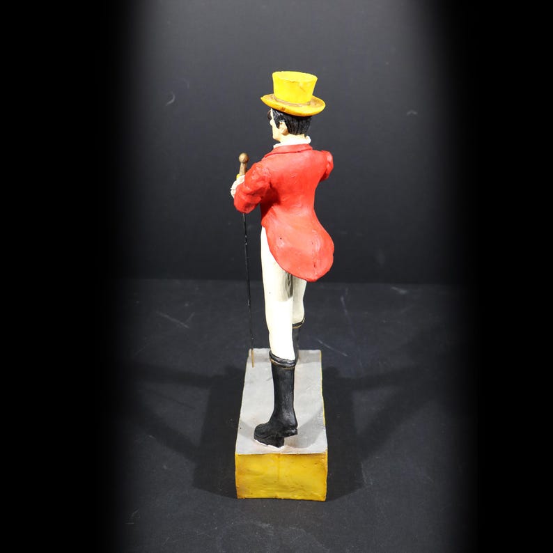 May include: A vintage figurine of a man in a red tailcoat, yellow top hat, and black boots, holding a cane. The figure stands on a rectangular base with a yellow bottom and grey top. The figurine is viewed from the back.